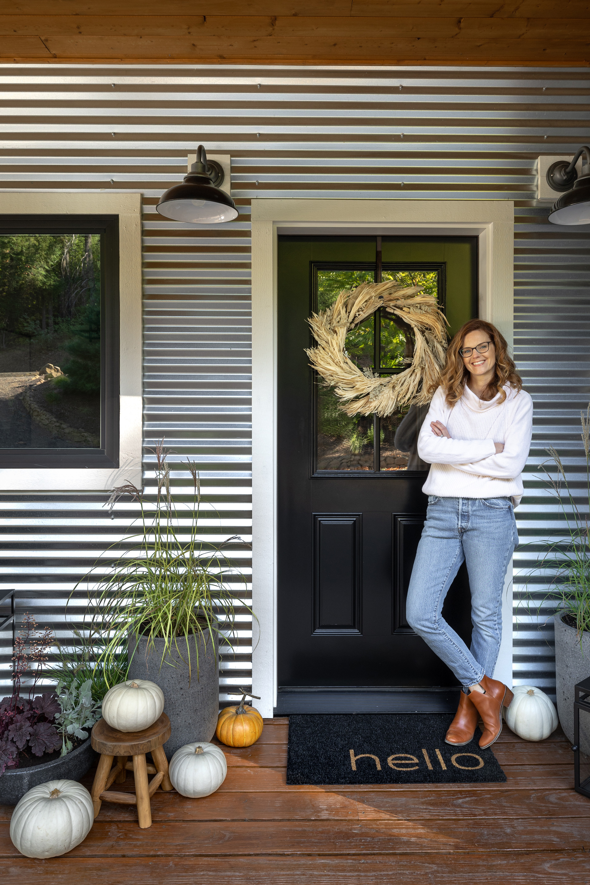 Ashley Hinkle standing with arms crossed and smiling in front of her black front door with fall decorations such as pumpkins, a wreath, and plants